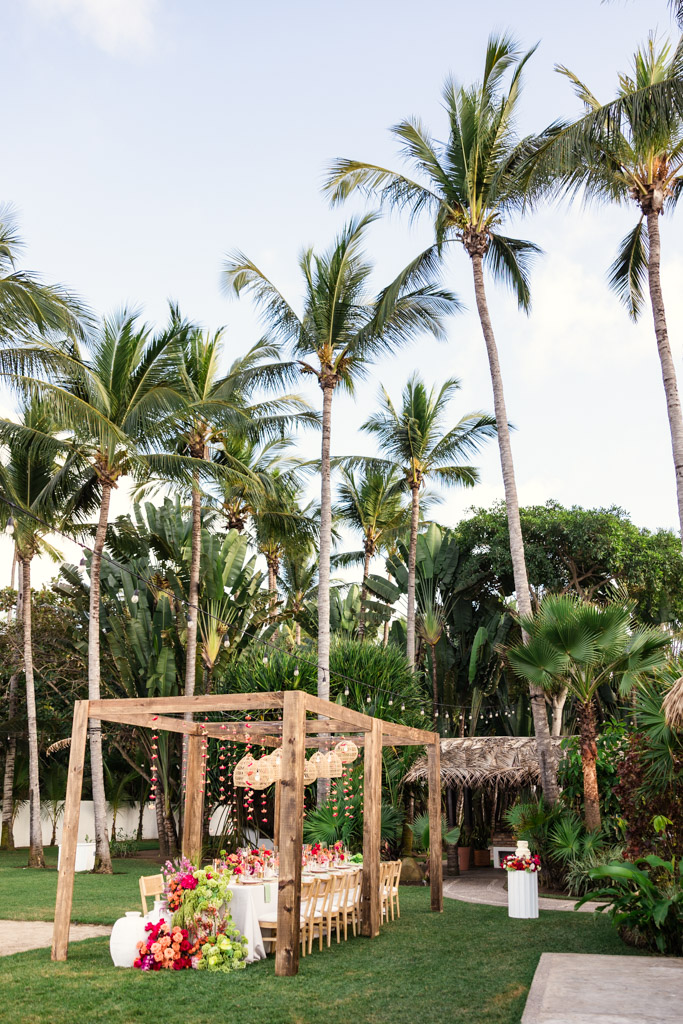 A decorated outdoor dining table under a wooden pergola, surrounded by palm trees and lush greenery, set for a Frente al Punto wedding celebration