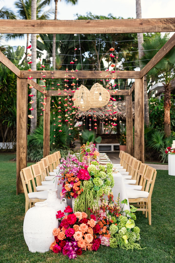 Outdoor table setup with wicker chairs, colorful floral arrangements, and hanging lights under a wooden pergola for Frente al Punto wedding