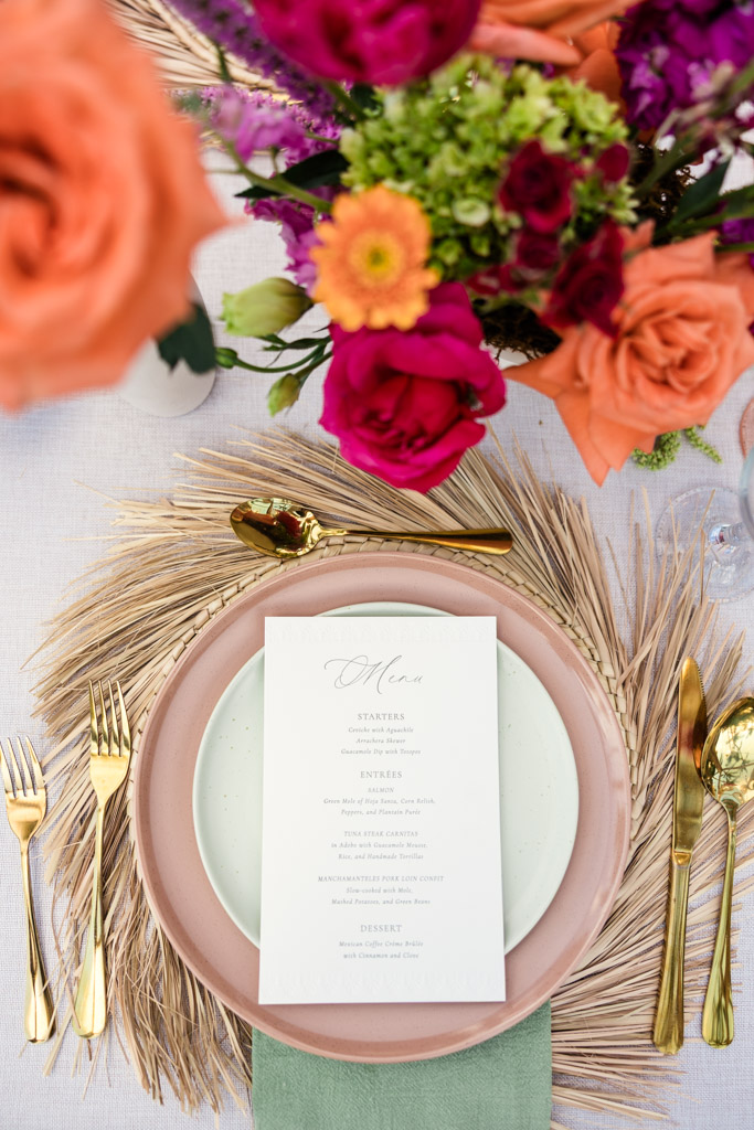 Guest table setting with a menu, gold cutlery, and colorful flowers on a woven placemat for a Frente al Punto wedding celebration