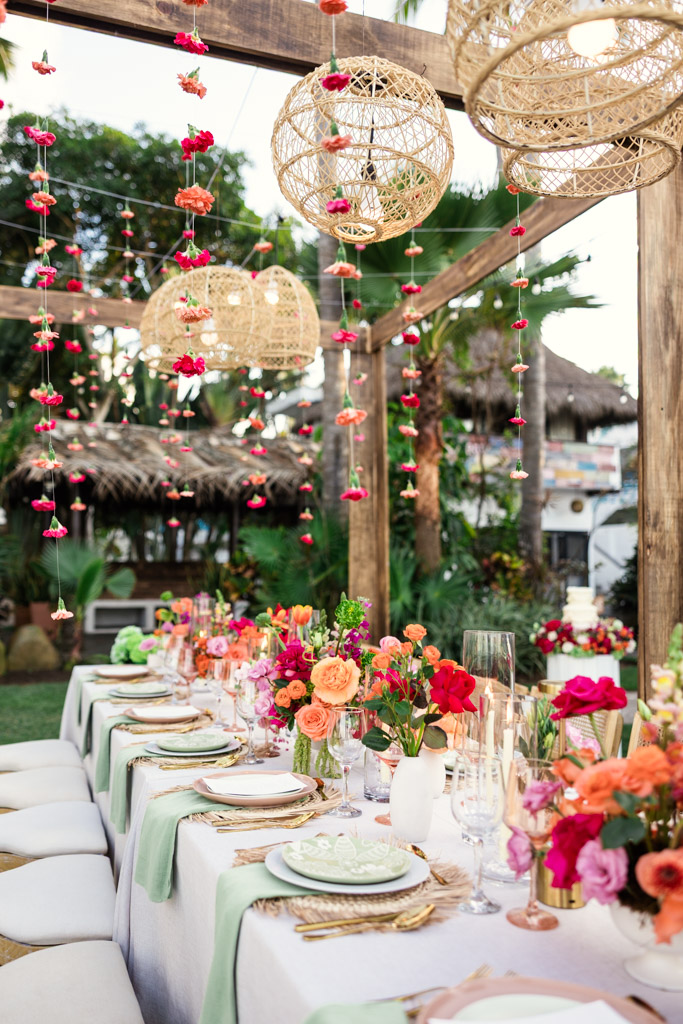 A long outdoor table is set with colorful flowers, glassware, and hanging lanterns in a tropical garden for a Frente al Punto wedding celebration