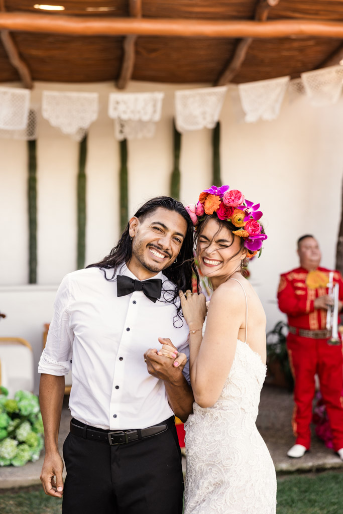 Smiling couple in wedding attire at a Frente al Punto wedding, bride with a colorful flower crown, and a mariachi musician in the background