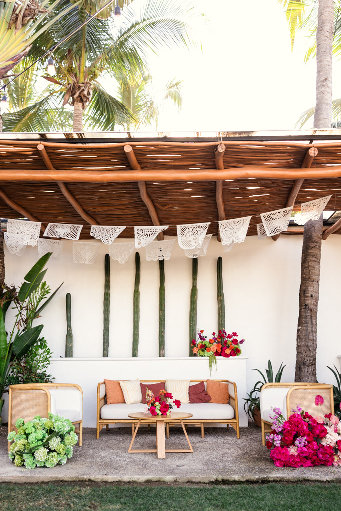 Outdoor patio with bamboo roof, white couches, colorful flowers, and hanging papel picado decorations for a Frente al Punto wedding celebration
