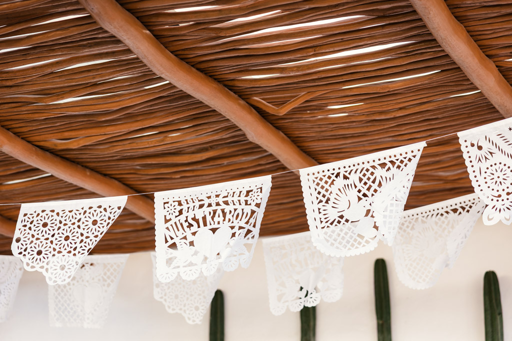 White papel picado banners hang under a woven wooden ceiling, with cacti visible below for Frente al Punto wedding