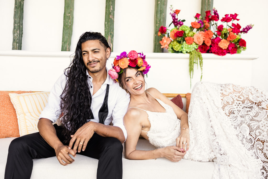 Smiling couple in wedding attire sits on a couch with bright floral arrangements in the background, during their Frente al Punto wedding