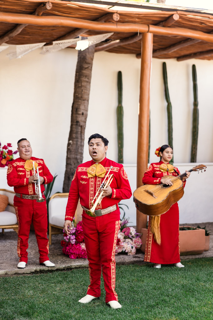 Three mariachi musicians in red suits perform outdoors at a Frente al Punto wedding; one sings, another plays a trumpet, and one holds a guitar