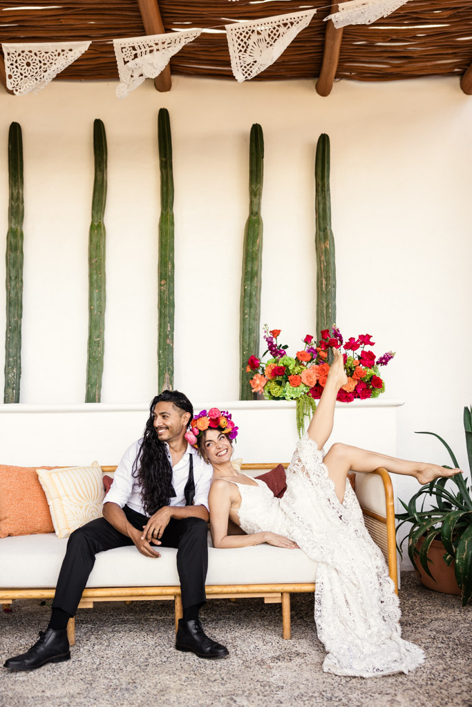 Bride and groom sit on a couch, smiling, with colorful flowers and tall cacti in the background at their Frente al Punto wedding