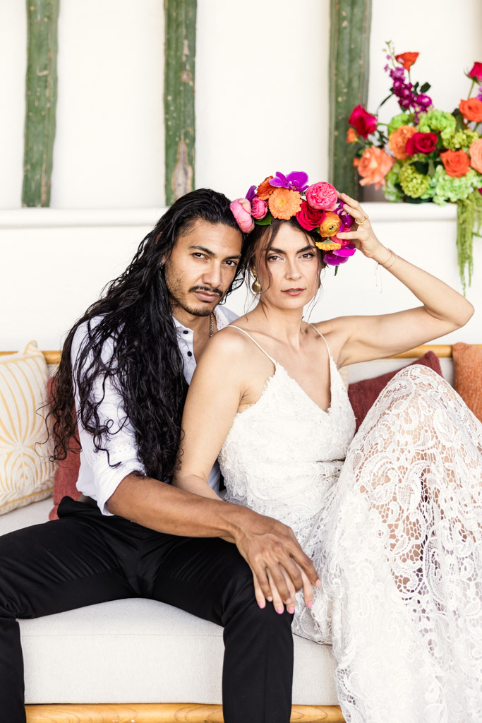 A couple sits on a couch at their Frente al Punto wedding; the woman wears a lace dress and floral crown, while the man has long dark hair