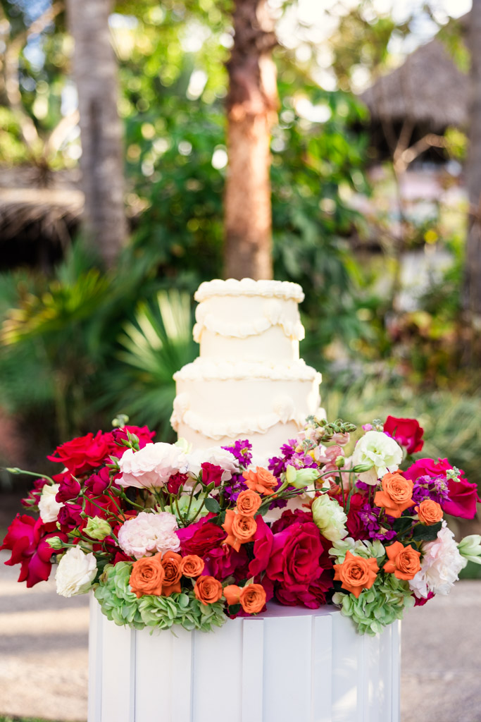 A white tiered cake is displayed outdoors at a Frente al Punto wedding, surrounded by vibrant red, orange, and pink flowers