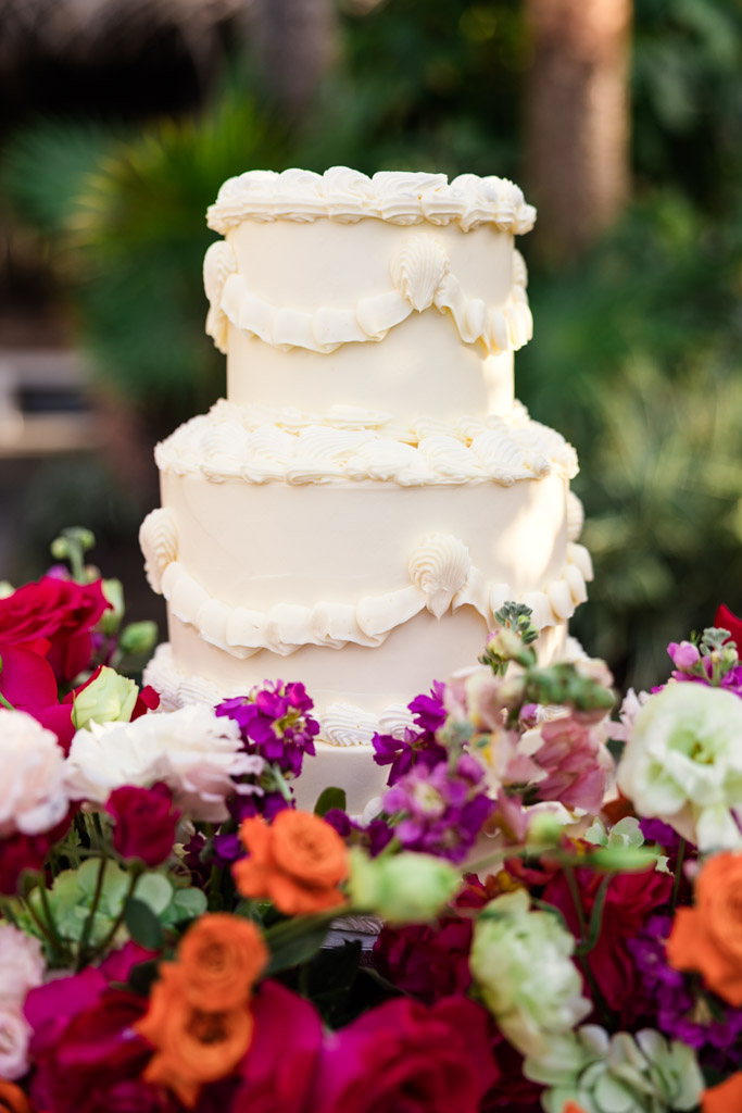 Three-tier white frosted cake surrounded by colorful flowers, with greenery in the background for a Frente al Punto wedding