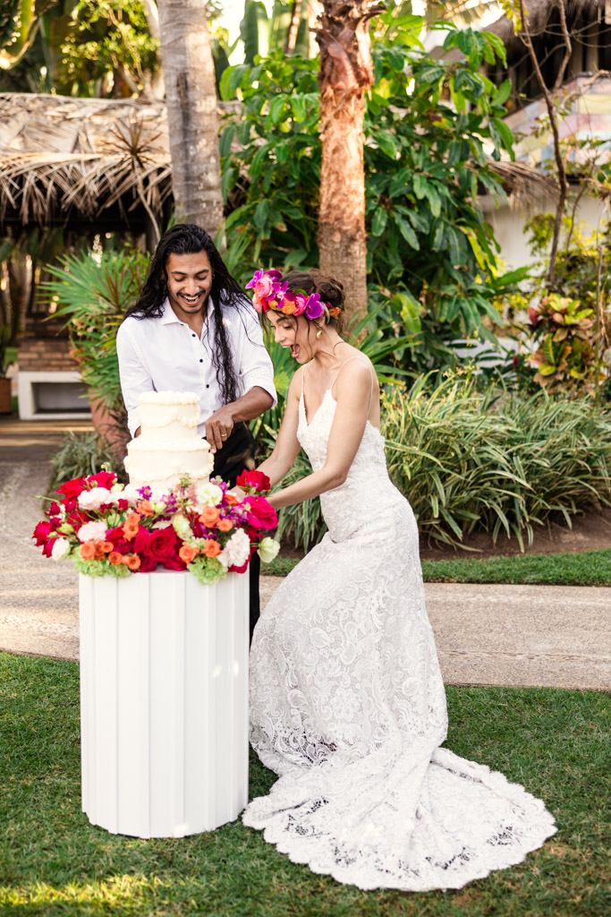 Bride and groom smiling as they cut a white wedding cake outdoors, surrounded by colorful flowers at their Frente al Punto wedding in Sayulita