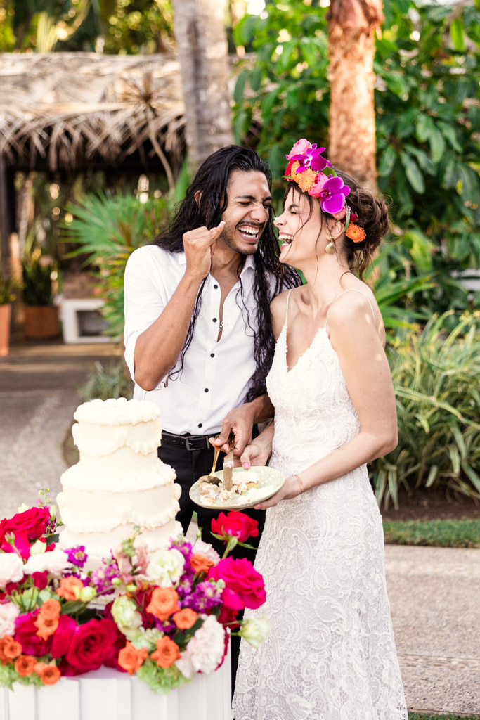 A joyful couple in wedding attire laughs while cutting cake outdoors, surrounded by colorful flowers at their Frente al Punto wedding