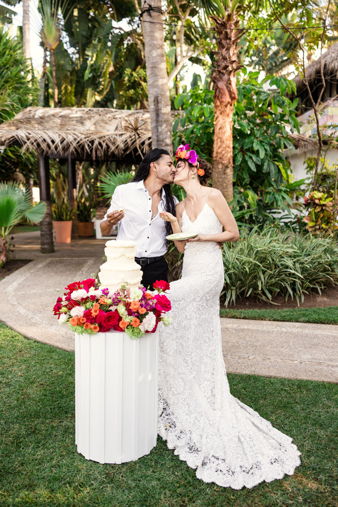 Bride and groom smile and kiss by a wedding cake surrounded by bright flowers in a lush outdoor setting at their Frente al Punto wedding
