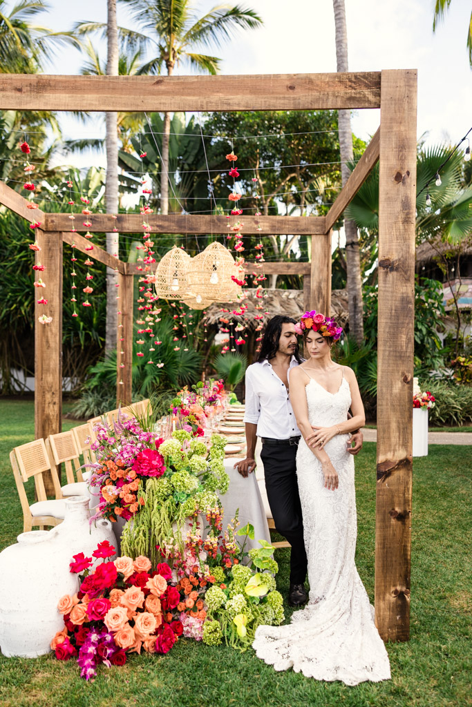 A bride and groom pose by a vibrant, flower-filled outdoor wedding table under a wooden pergola at their Frente al Punto wedding