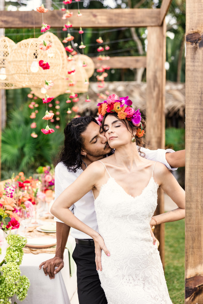 A groom kisses the bride’s cheek as she smiles, both surrounded by vibrant flowers at their Frente al Punto wedding