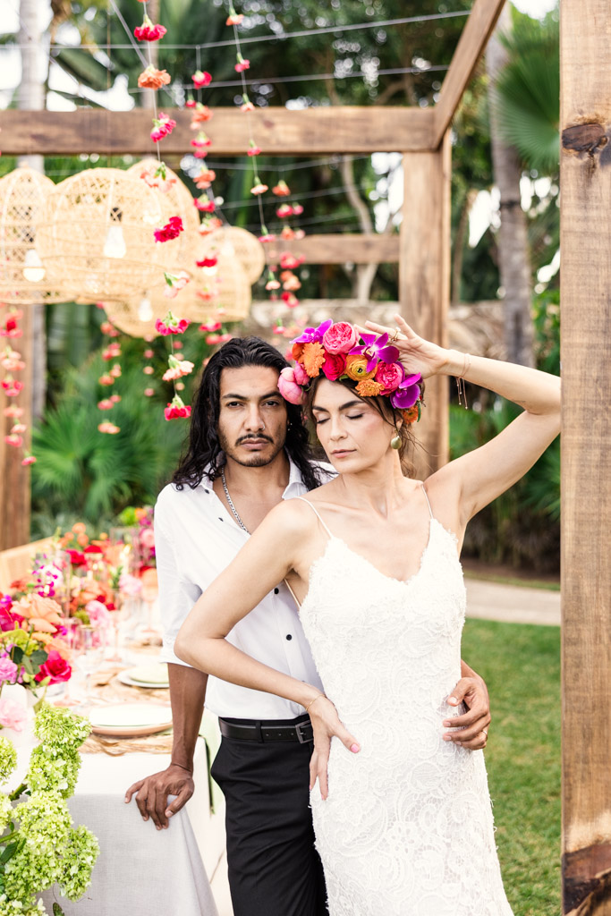A couple poses by a garden table at their Frente al Punto wedding; the woman wears a white dress and a bright flower crown