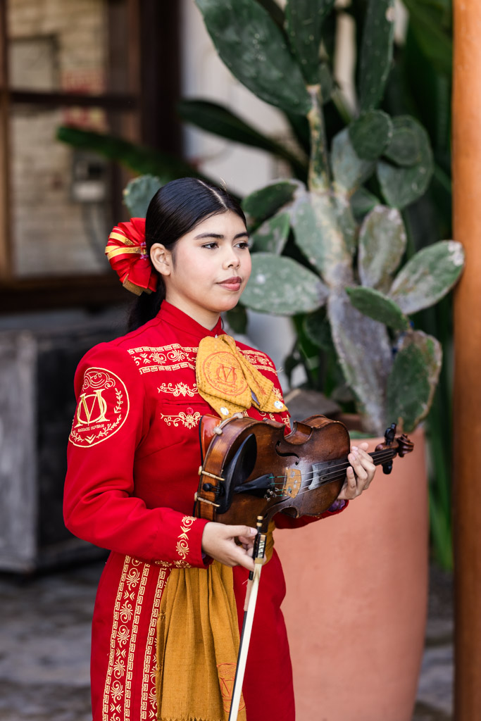 A woman in a red mariachi outfit holds a violin, standing in front of a large potted cactus plant, for Frente al Punto wedding