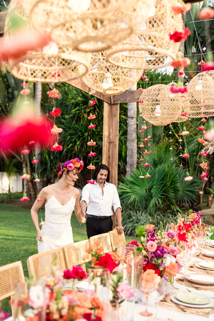 A bride and groom smile as they walk by a colorful, flower-filled Frente al Punto wedding table under hanging lanterns