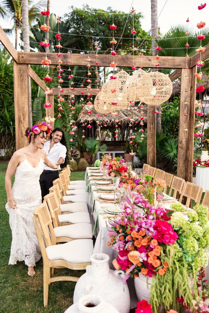 Bride and groom walk by a vibrant outdoor Frente al Punto wedding table decorated with colorful flowers and hanging lanterns