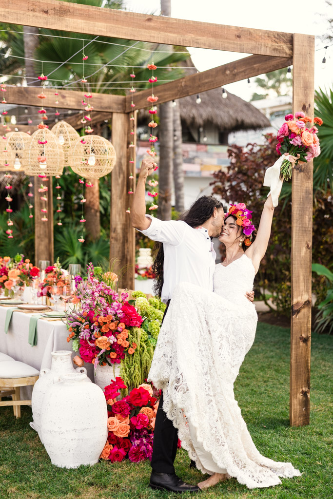 At a Frente al Punto wedding, a groom lifts and kisses his bride holding a bouquet, surrounded by vibrant flowers at an outdoor celebration