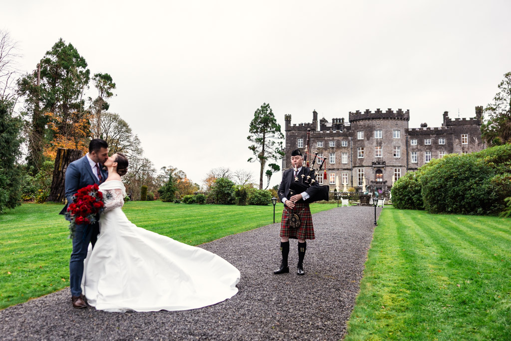 A bride and groom kiss on a path as a bagpiper plays, with Markree Castle in the background