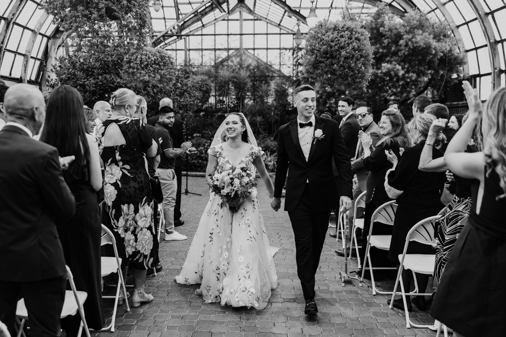Black and white photo of bride and groom walking down the aisle smiling as guests applaud after their Lincoln Park Conservatory wedding ceremony