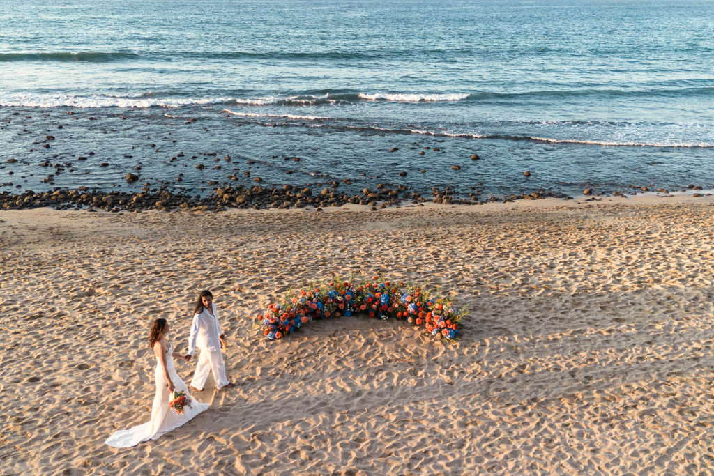 Couple in white walks on the beach in Sayulita near a colorful flower arrangement, with the ocean in the background