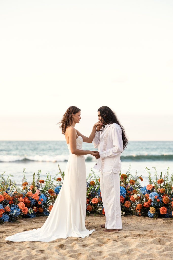 Bride and groom stand on the beach holding hands, surrounded by colorful flowers, with the ocean behind them during their Sayulita beach elopement
