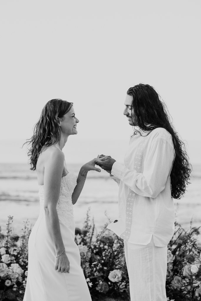 Black and white photo of couple in white standing by the ocean, exchanging rings at an outdoor wedding ceremony, smiling at each other during their Sayulita beach elopement