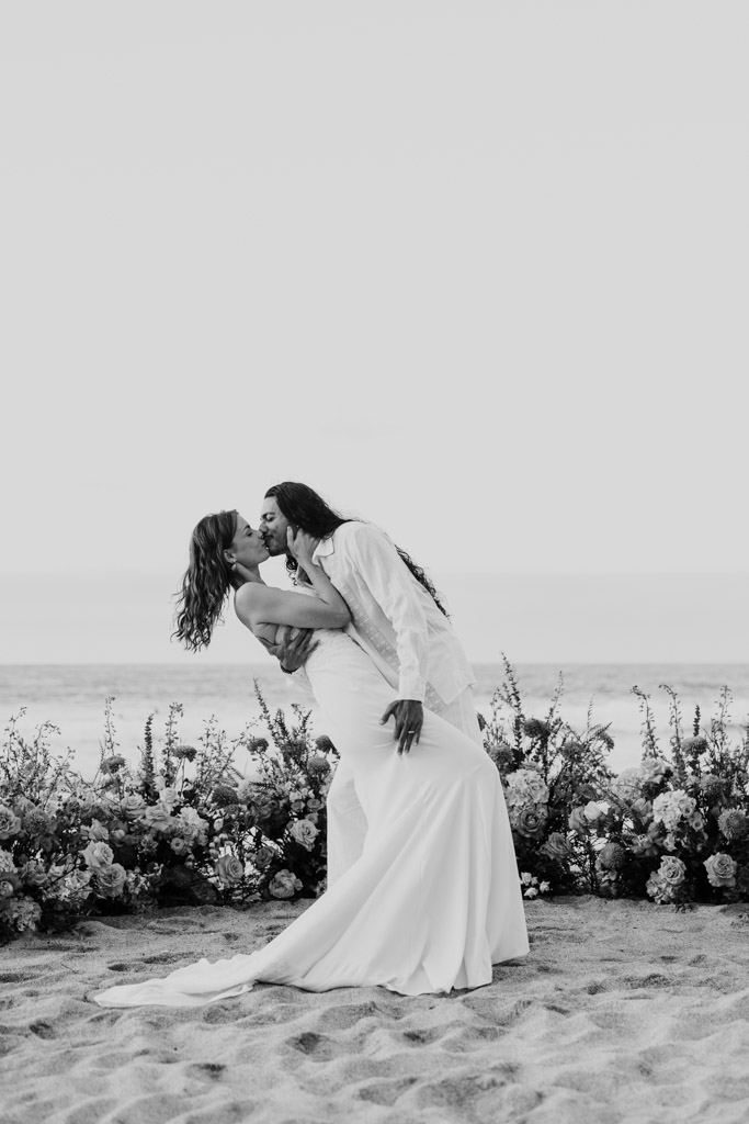 Black and white photo of couple in wedding attire kissing during their Sayulita beach elopement, surrounded by flowers, with the ocean in the background