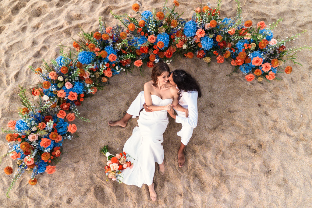 Bride and groom in white attire sit embracing on sand, surrounded by a semicircle of colorful flowers during their Mexico beach elopement