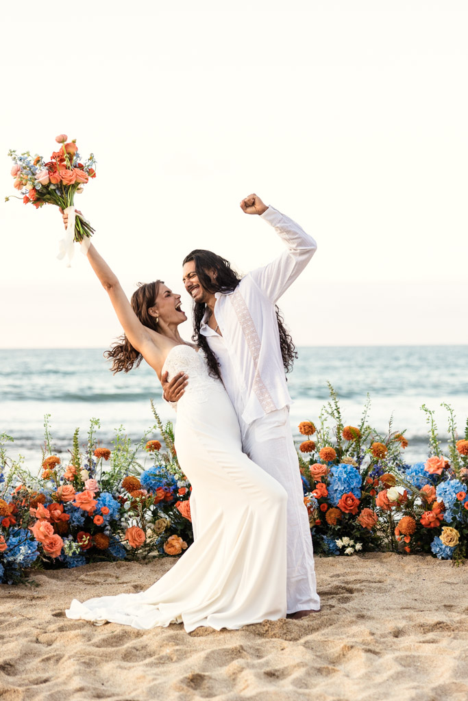 A joyful couple celebrates their Mexico beach elopement in wedding attire, surrounded by colorful flowers