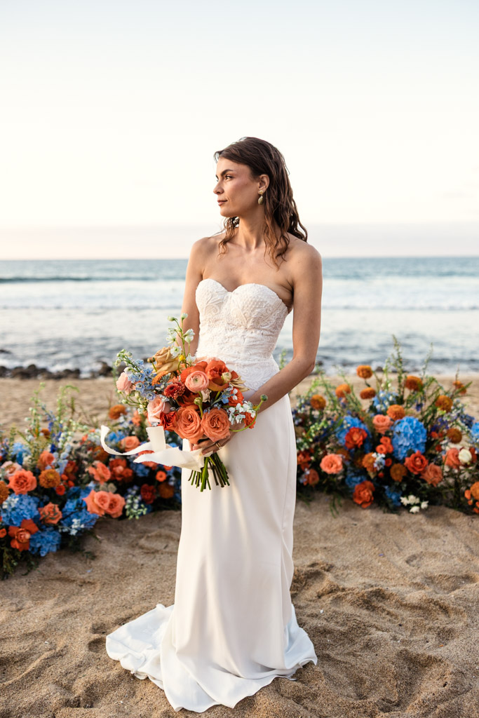 Bride in a strapless white gown holding a bouquet, standing on a Mexico beach with colorful flowers behind her