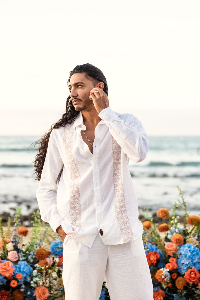 Portrait of groom in white clothing standing on the beach, touching his ear, with colorful flowers in the background