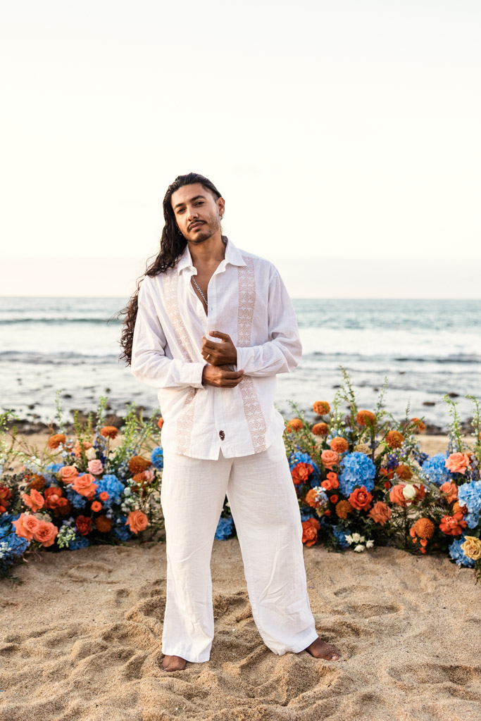 Groom in white outfit stands barefoot on sandy beach during his Mexico beach elopement, with colorful flowers and the ocean in the background