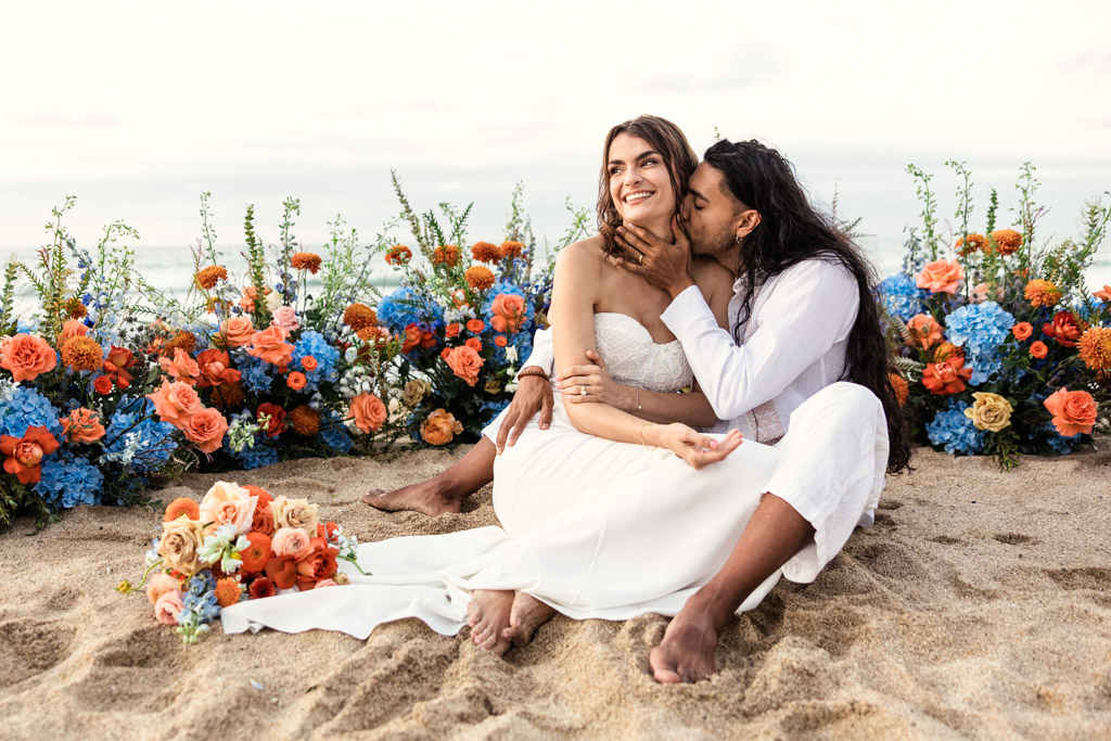 A couple sits on a sandy Mexico beach, surrounded by colorful flowers, smiling and embracing during their elopement