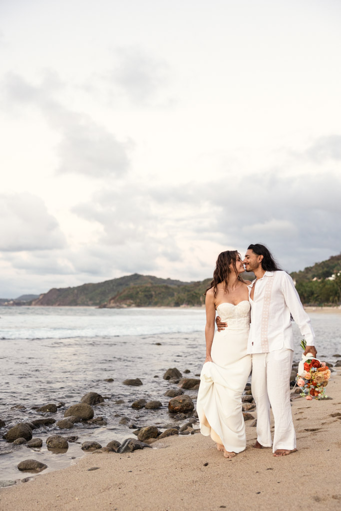 A couple in wedding attire walks barefoot on a Mexico beach, smiling at each other, with the ocean and hills behind them during their Sayulita beach elopement
