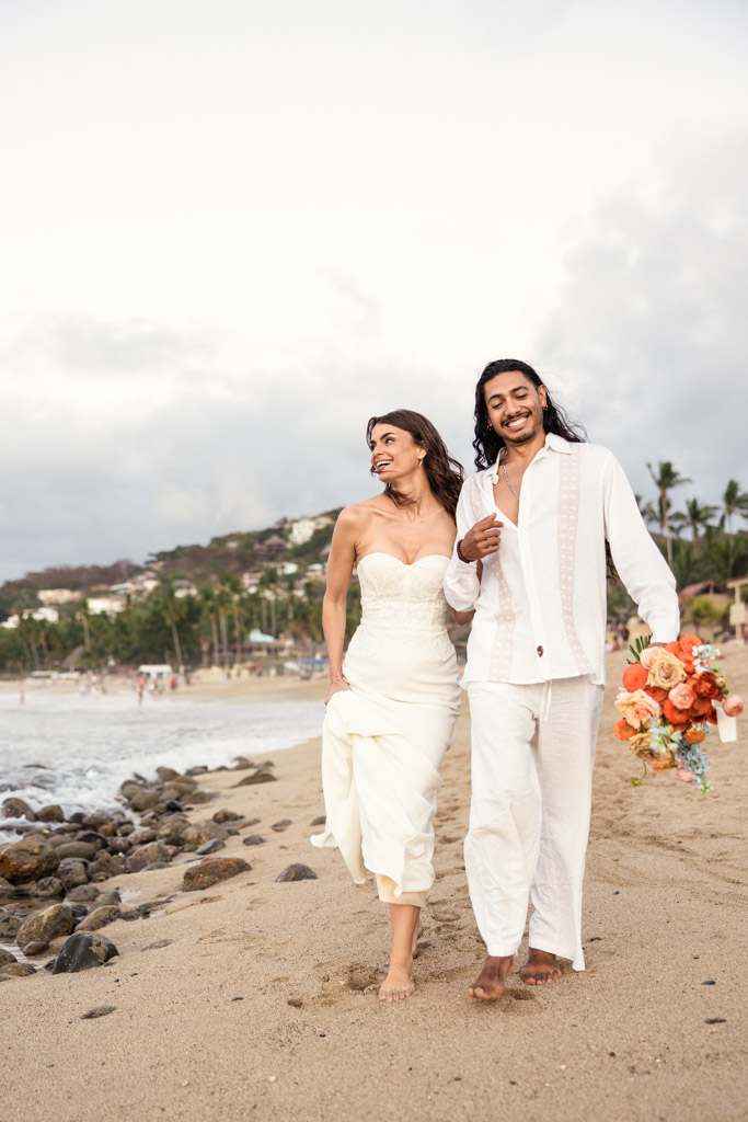 A couple in white attire walks barefoot on a sandy beach during their Mexico beach elopement, smiling and holding a bouquet of orange flowers