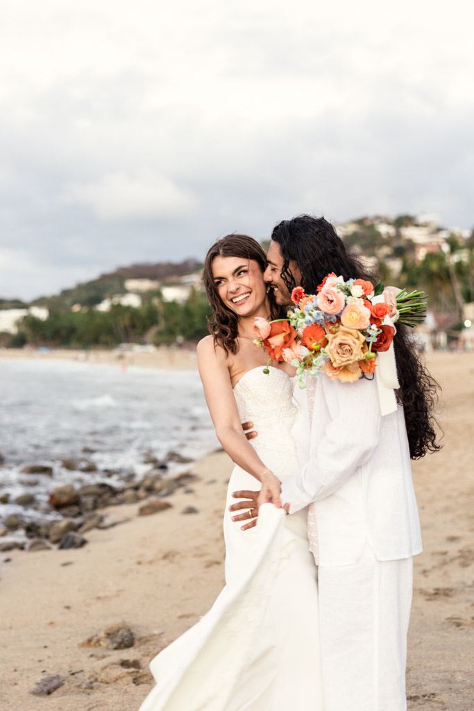 Bride and groom smiling and embracing on a sandy beach during their Mexico beach elopement, with the bride holding a colorful bouquet of flowers