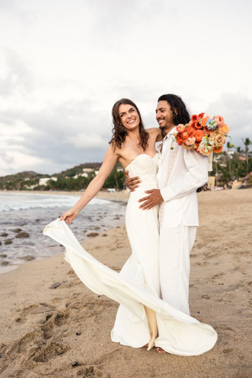 Smiling bride and groom in white attire embrace on a sandy shore during their Mexico beach elopement, holding a colorful bouquet