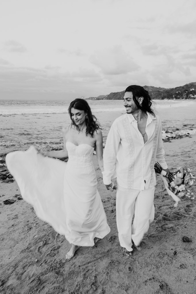Black and white photo of bride and groom in white outfits walking barefoot on a Mexico beach, smiling and holding hands during their elopement in Sayulita