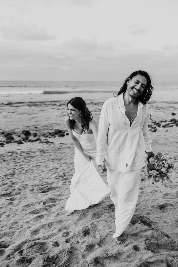 Black and white photo of happy couple in white attire walking barefoot on a sandy shore, smiling and laughing together during their Mexico beach elopement in Sayulita