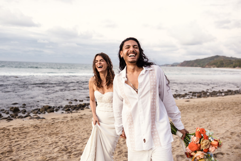 A joyful couple in wedding attire laughs together on a sandy shore, the groom holding a bouquet of flowers during their Mexico beach elopement