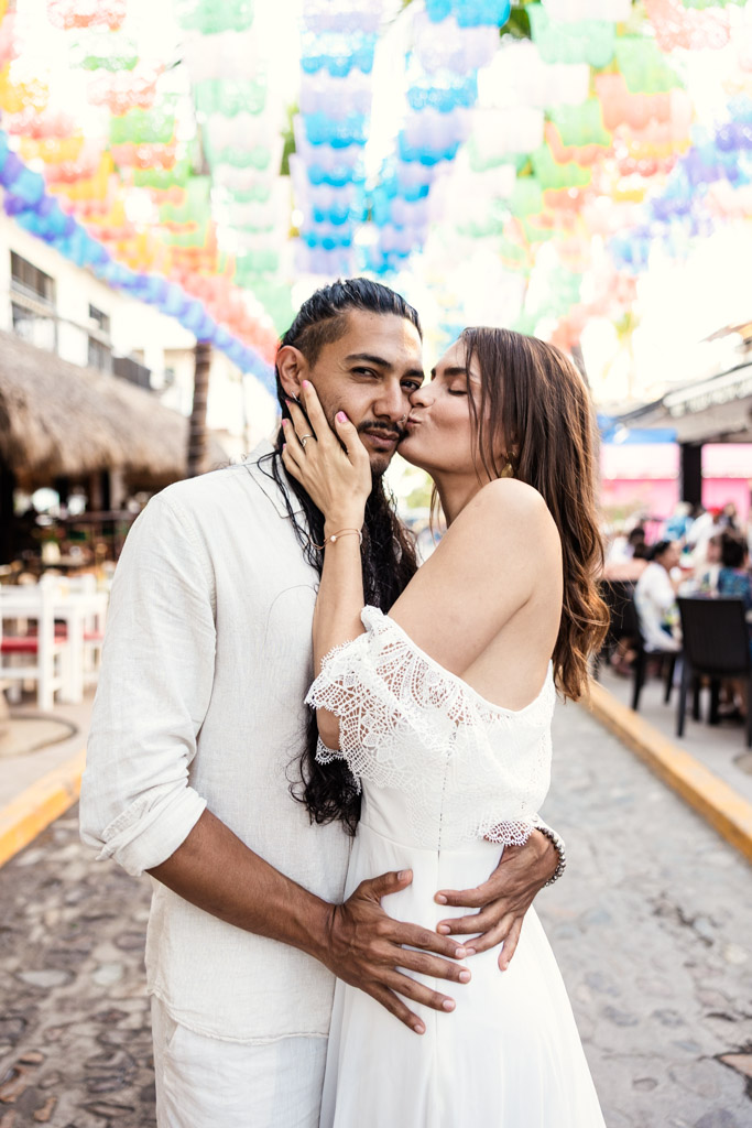 A couple in white outfits embrace and kiss on a colorful, decorated street outdoors during their Sayulita engagement session