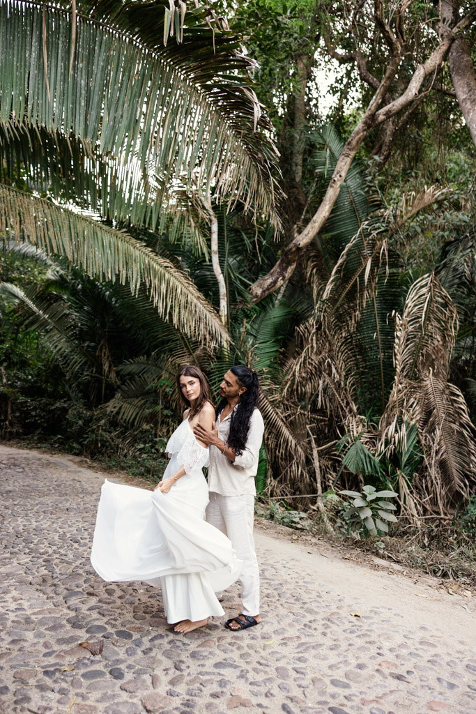 A couple in white outfits poses on a cobblestone path surrounded by lush tropical greenery during their Sayulita engagement session