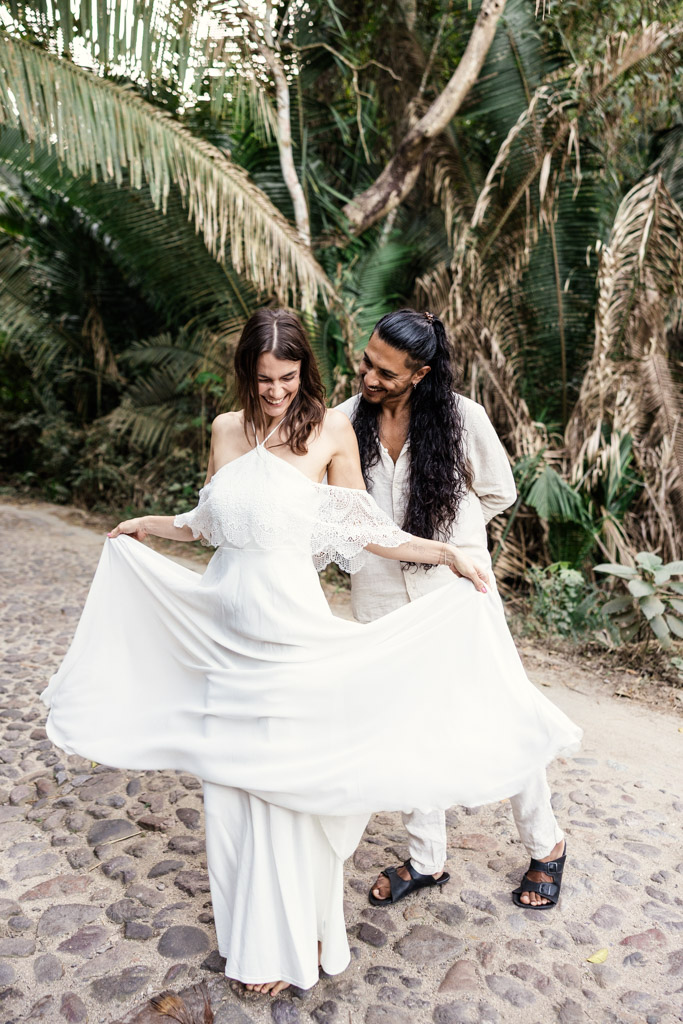 A smiling couple stands on a stone path in Sayulita; the woman holds out her white dress while the man looks at her