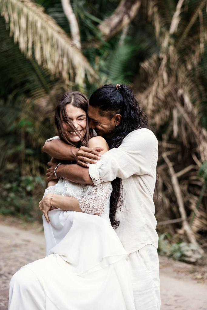 A couple in white clothes embraces joyfully outdoors, surrounded by lush tropical greenery, during their Sayulita engagement session