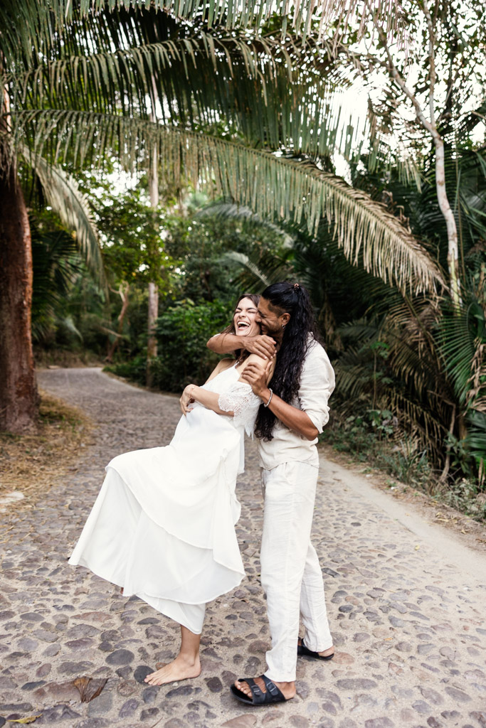 A joyful couple in white outfits laughs and embraces on a cobblestone path surrounded by lush greenery, during their Sayulita engagement session