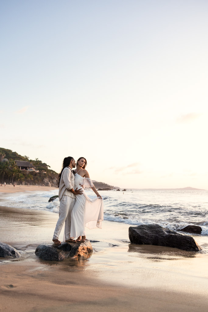 A couple in light clothing stands on rocks at a Sayulita beach as the sun sets by the ocean