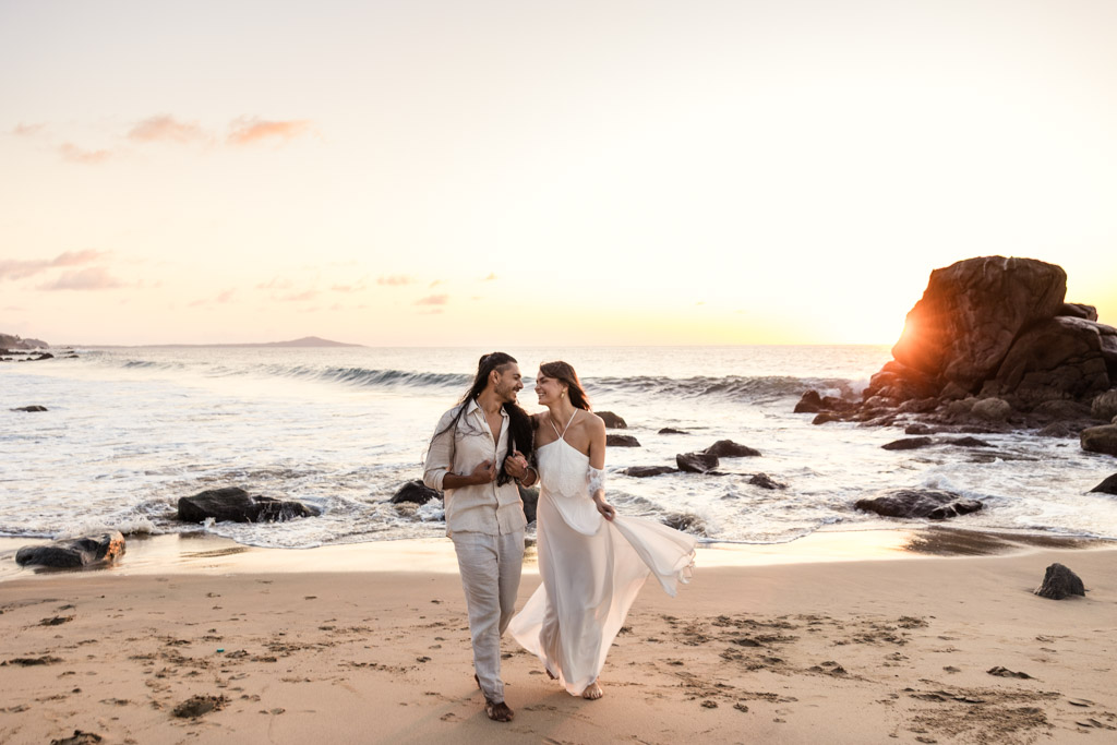 A couple walks hand in hand on a sandy beach at sunset, smiling at each other during their Sayulita engagement session