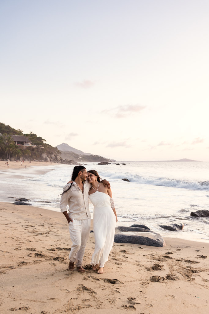 Couple dressed in white walks barefoot on a sandy beach at sunset during their Sayulita engagement session